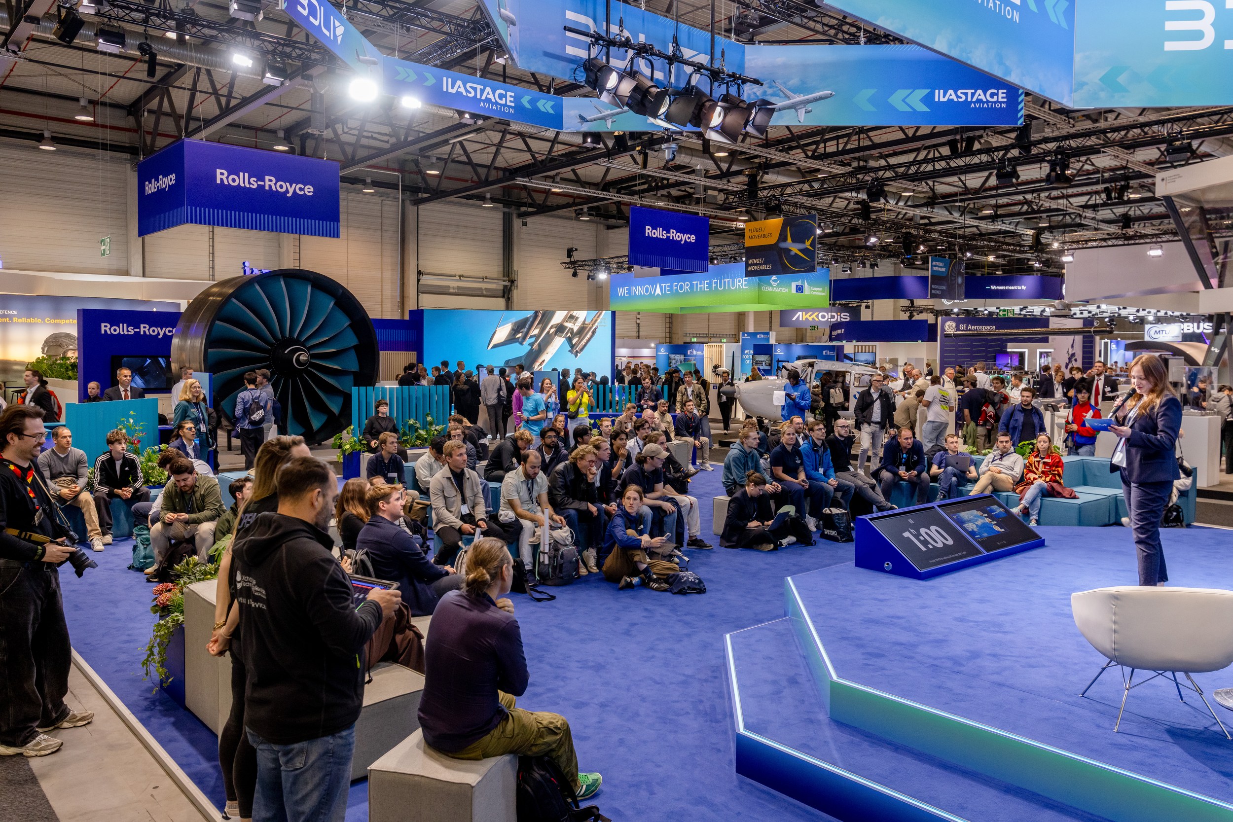 A moderator stands at the right edge of the ILA STAGE Aviation and speaks before a densely seated audience on low tiers.