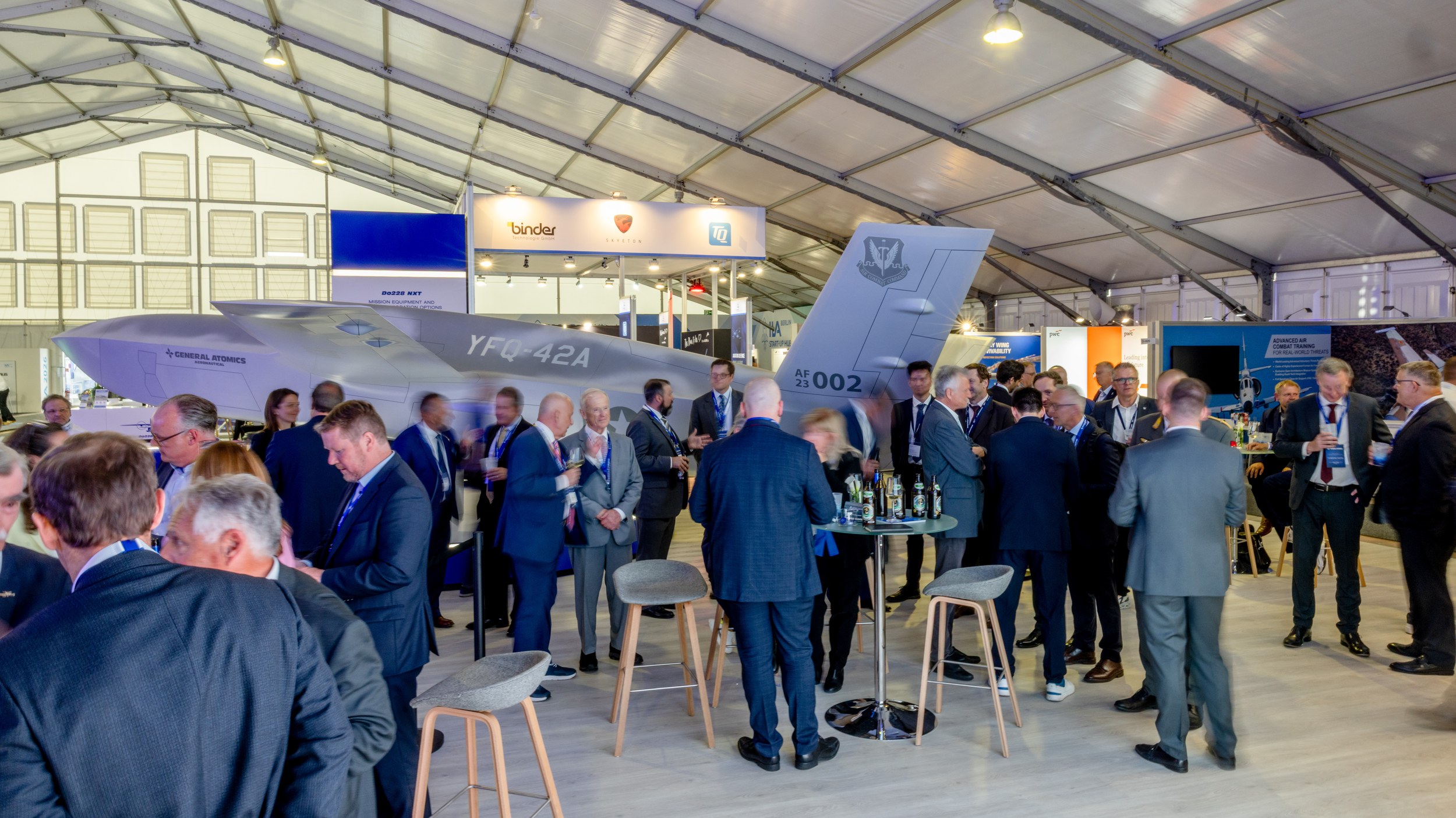 Numerous trade visitors gather in small conversation groups around high tables in front of a grey General Atomics …