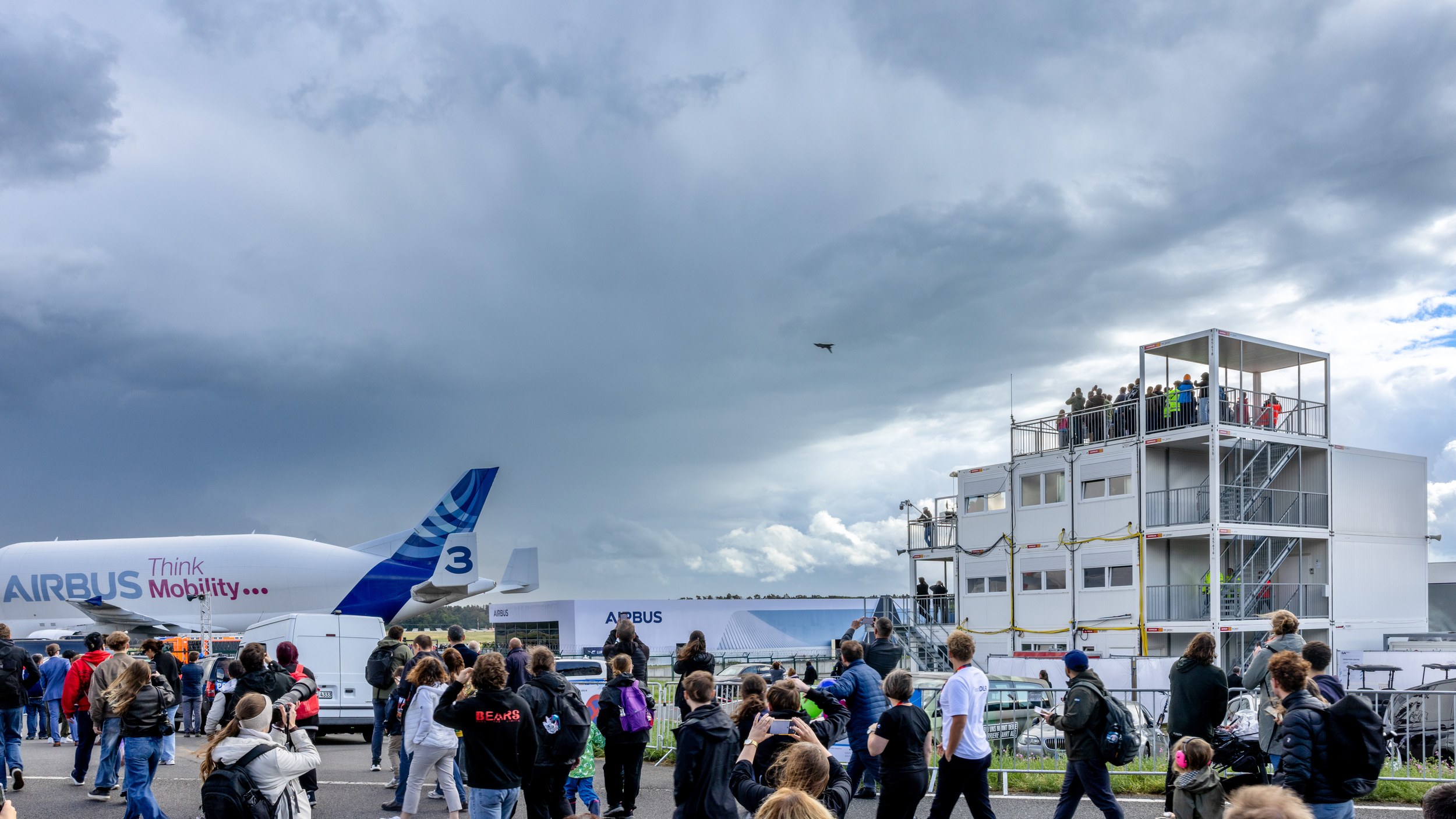 A dense crowd looks and photographs skyward as a small fighter jet flies against dark clouds.