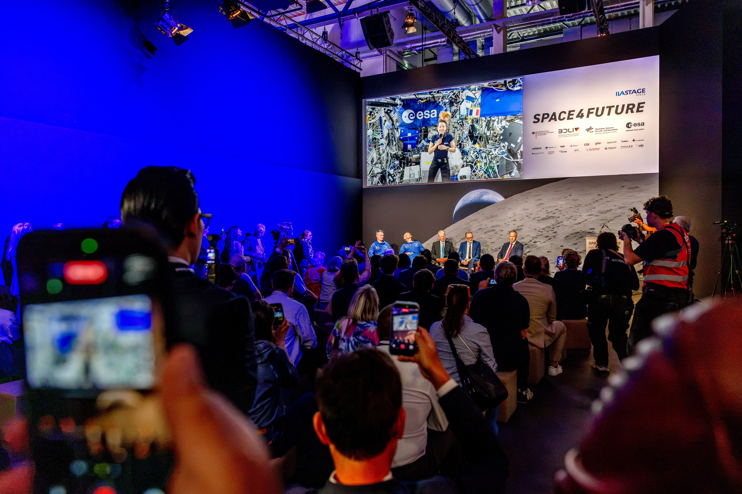 A dense audience watches and records a Space4Future panel on the ILA STAGE Space in front of a large lunar backdrop. Several guests sit on stage, including two people in blue suits, while the screen shows a person inside a space-station setting beside the esa logo.
