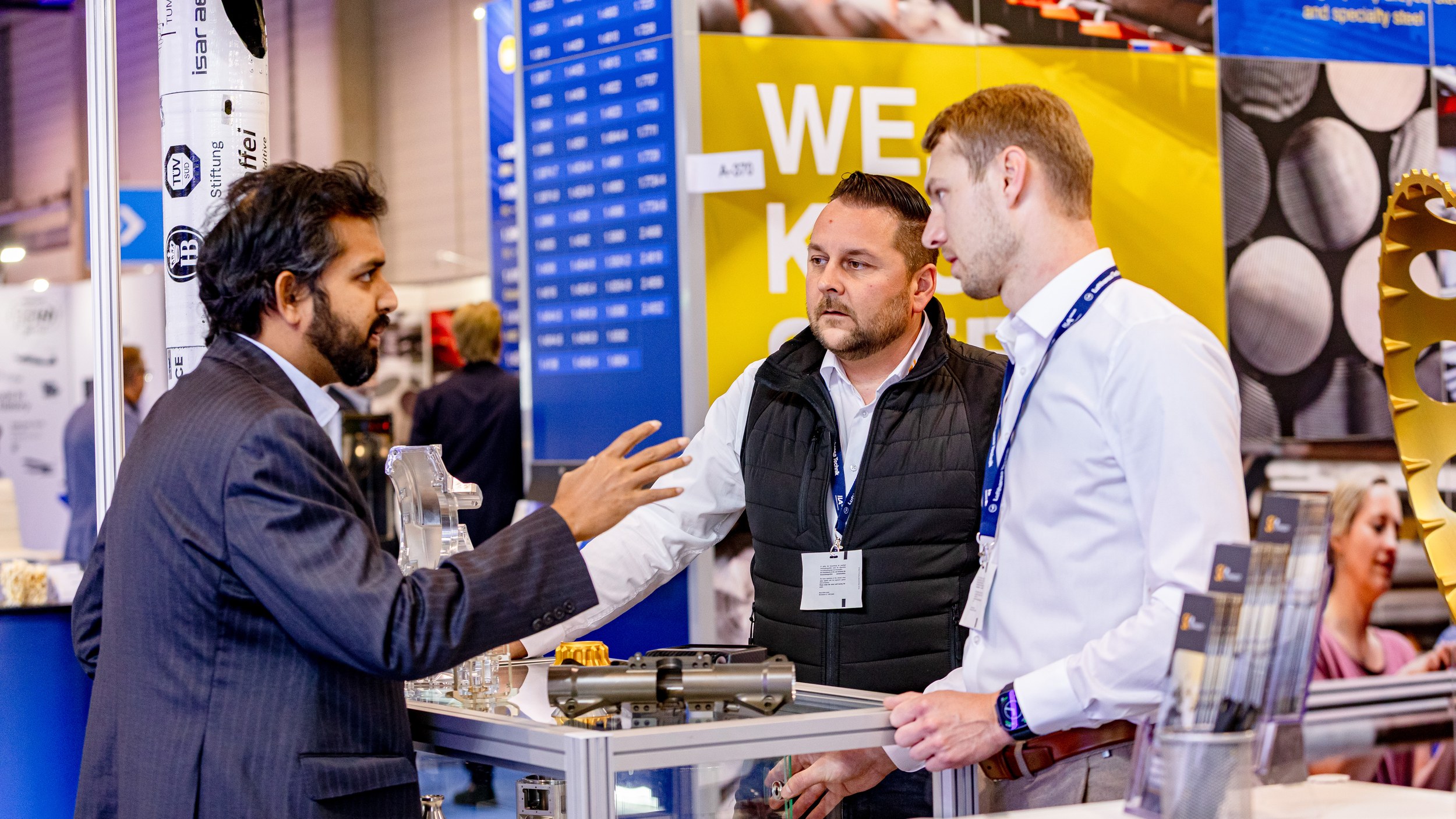 Three trade visitors discuss metal components displayed in a glass case, with the man on the left explaining something with his hand raised. Machined components and a gold-coloured gear structure sit on and beside the case, while TÜV SÜD marks are visible in the left background.