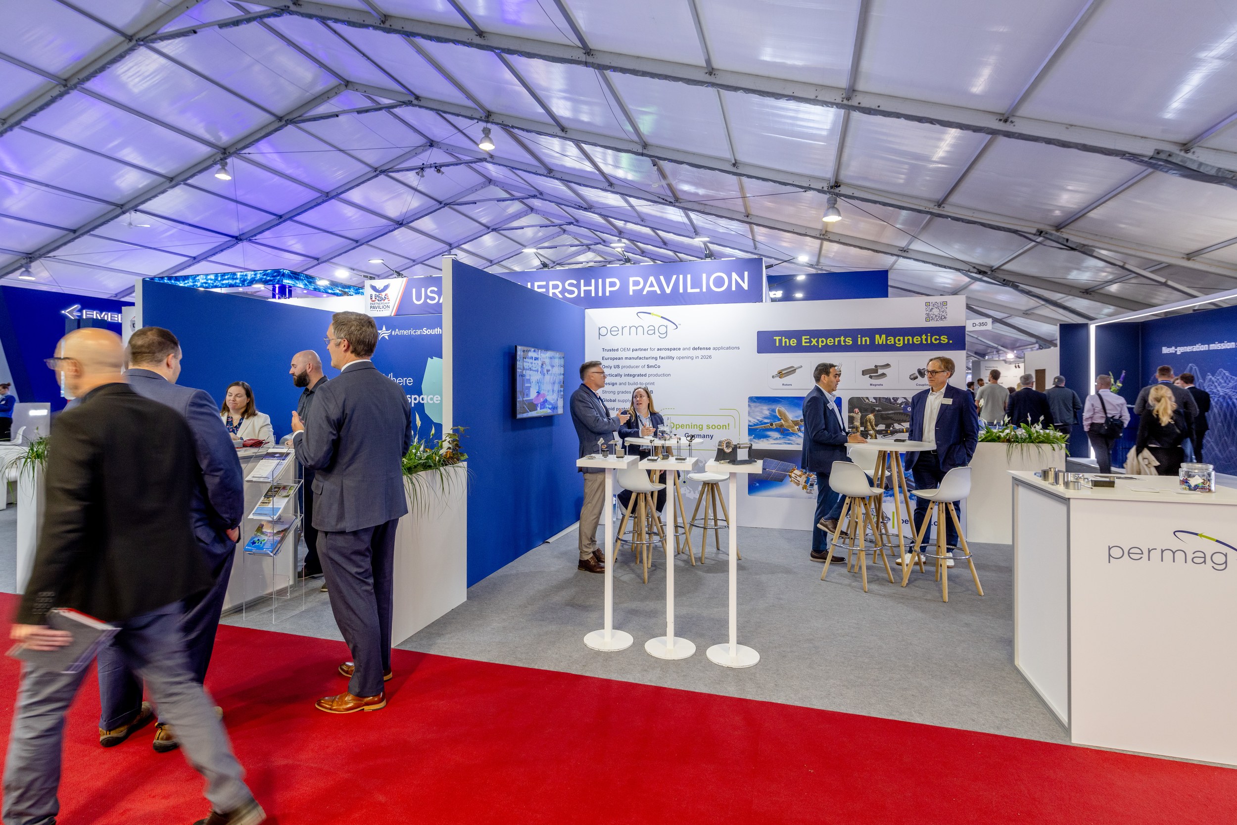 Inside the USA Partnership Pavilion in Hall D: trade visitors at high tables in front of blue-and-white stand walls along the red aisle.