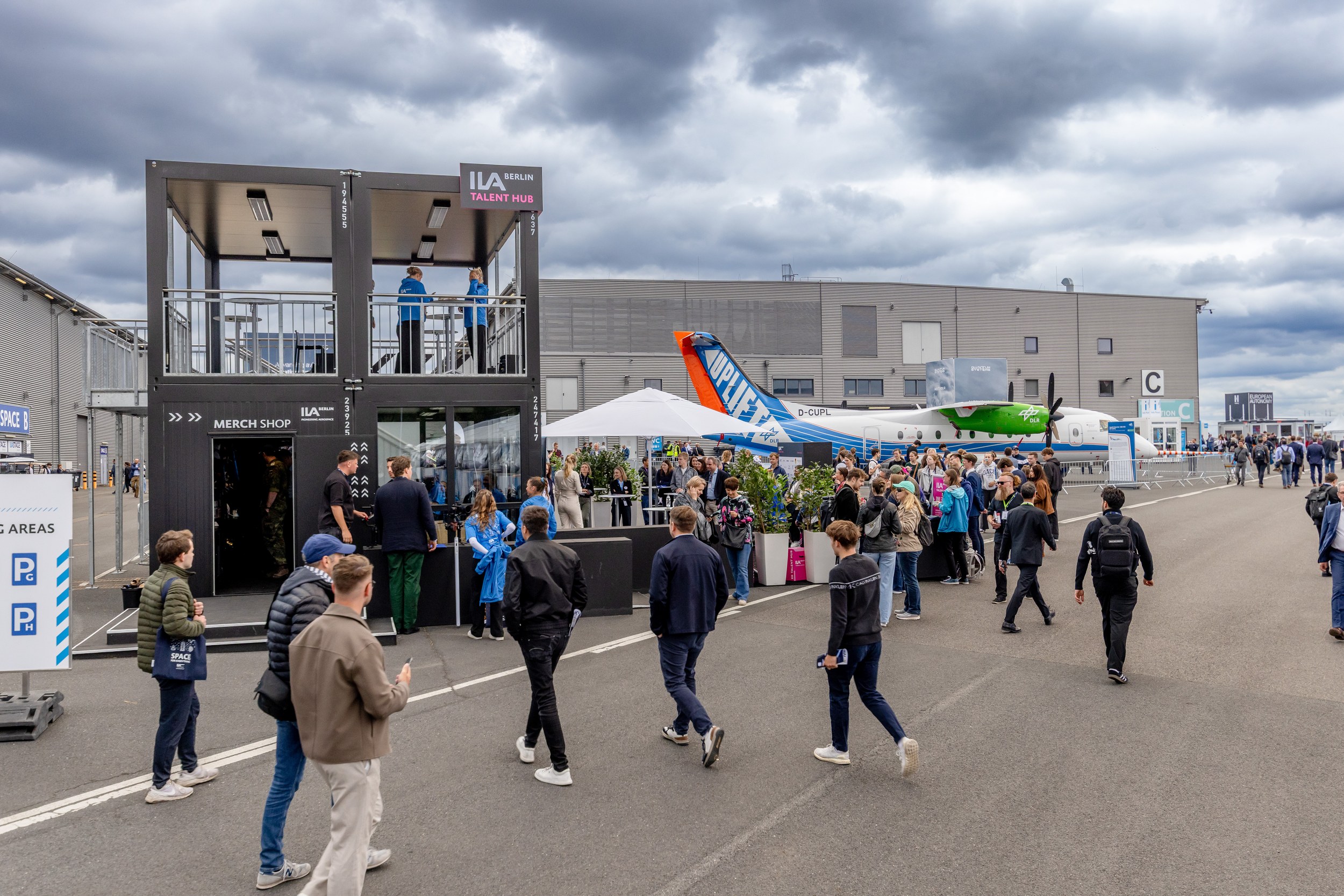 A large group of mostly young visitors gathers on the plaza in front of the two-level ILA Berlin Talent Hub and its merc