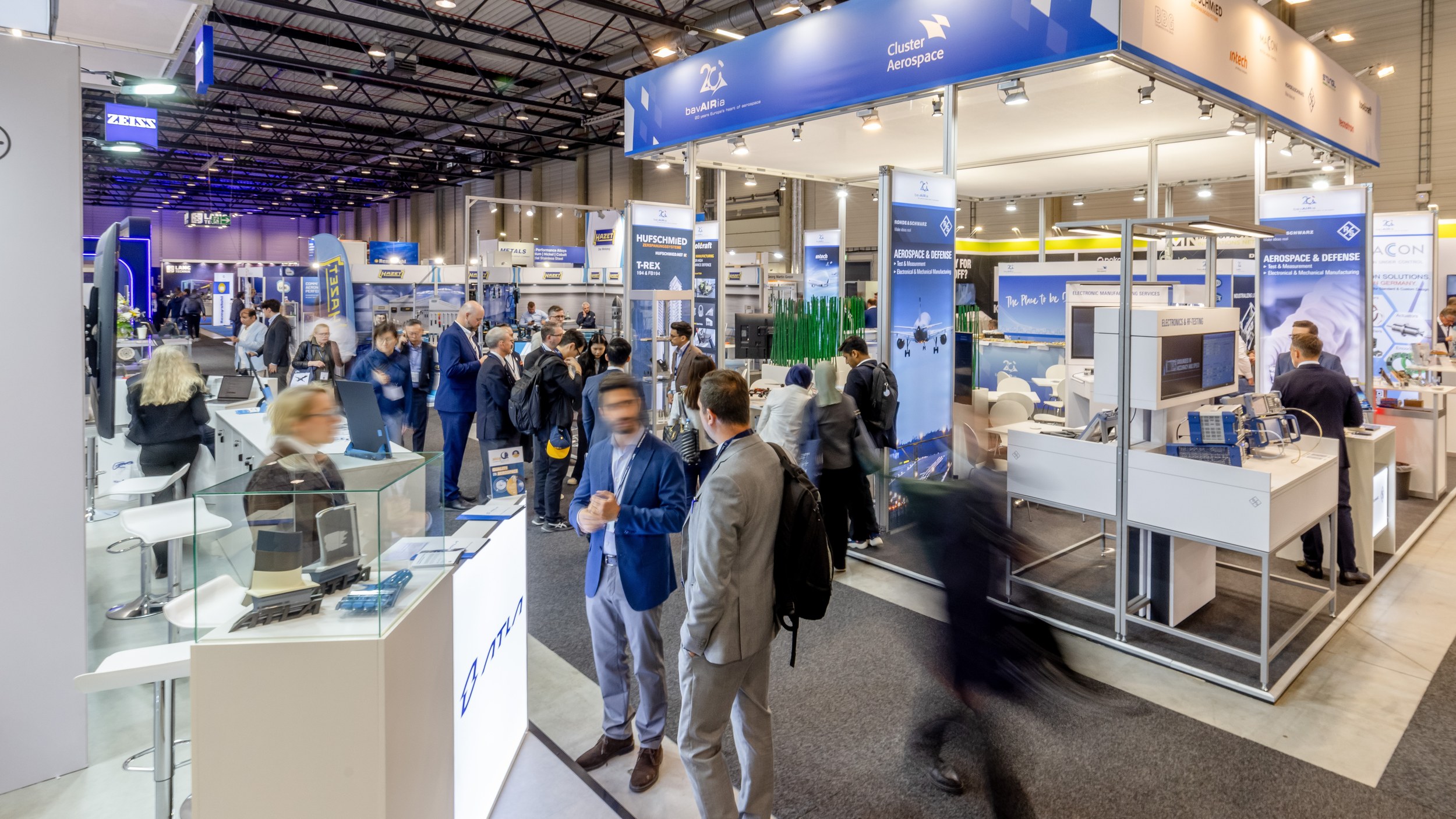 At the bavAIRia and Cluster Aerospace joint booth in Hall A, numerous trade visitors talk among display cases, counters 