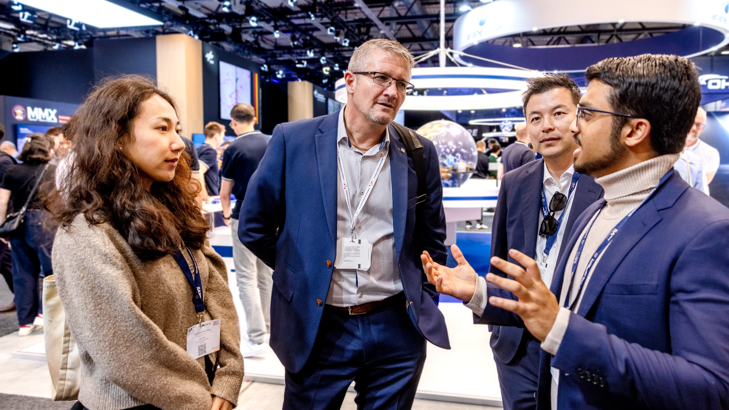 Four trade visitors stand in close conversation in front of a space exhibit island in Hall B, one explaining with both hands.