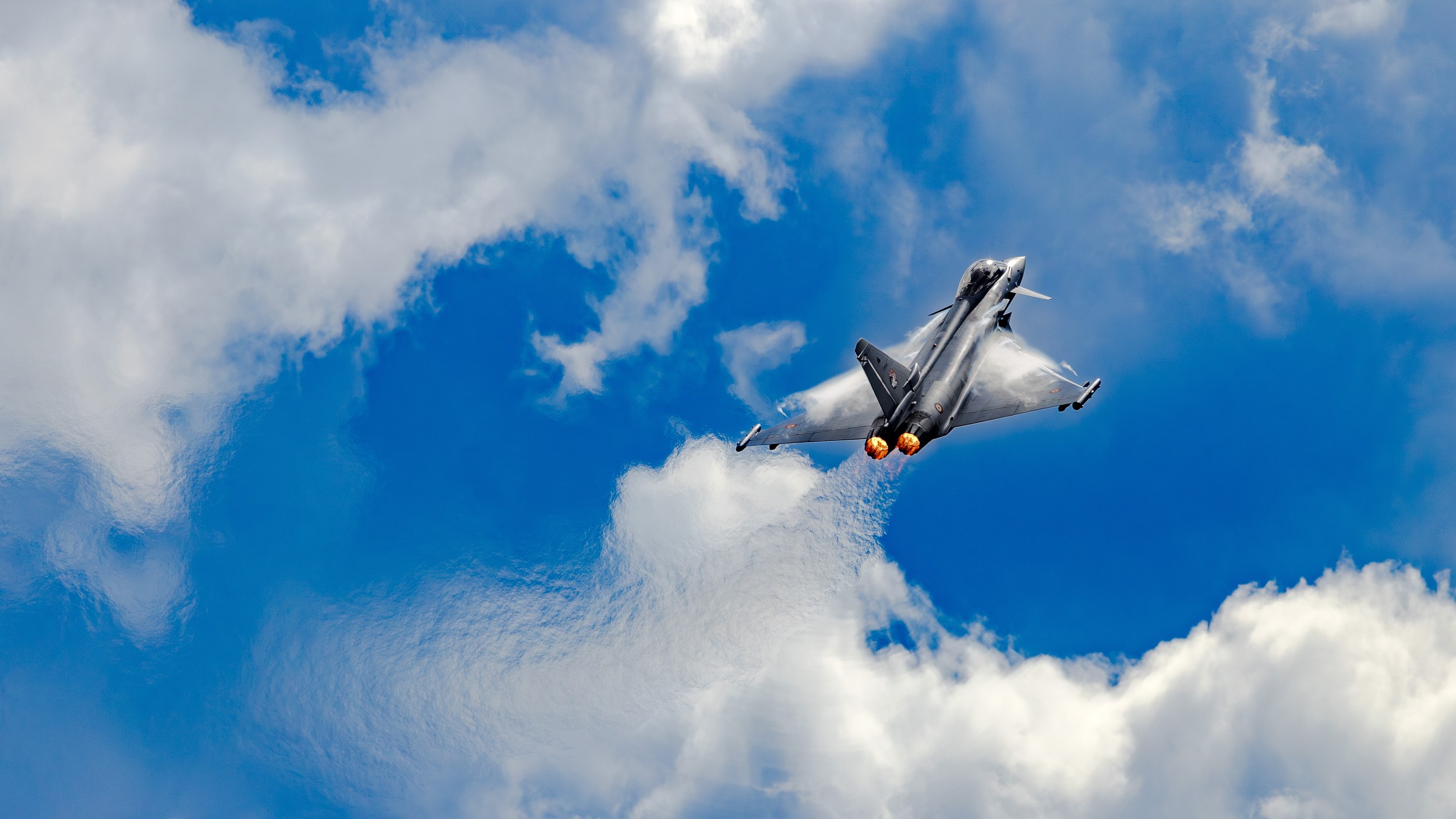A Eurofighter Typhoon climbs away from the camera during the Flying Display with both afterburners glowing bright orange. Broad condensation clouds form over the wings, followed by a textured vapor trail against blue sky and white clouds.