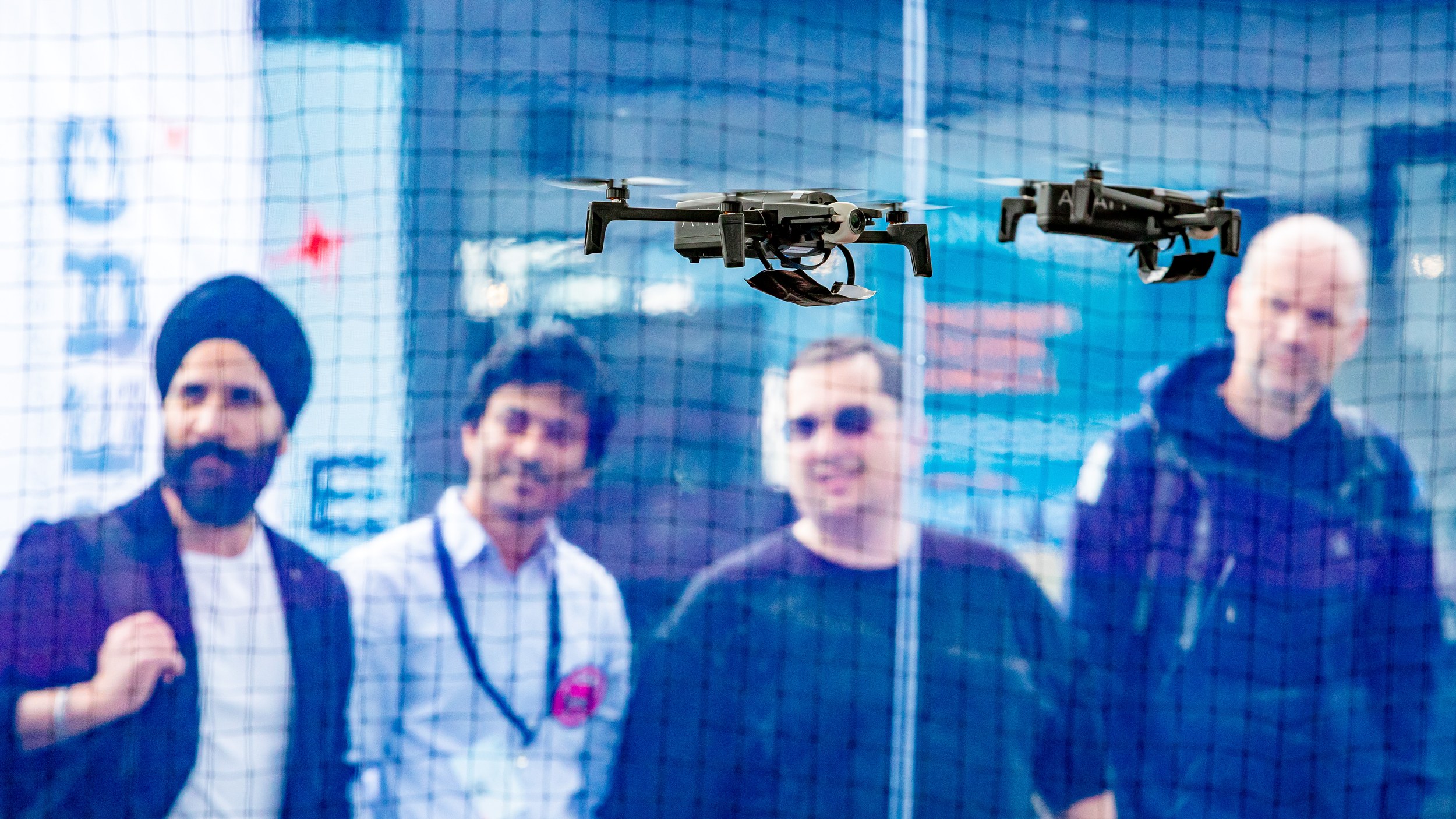 Two small quadcopters hover side by side behind the safety net in the Drone Pavilion at ILA Berlin 2026. Four visitors w