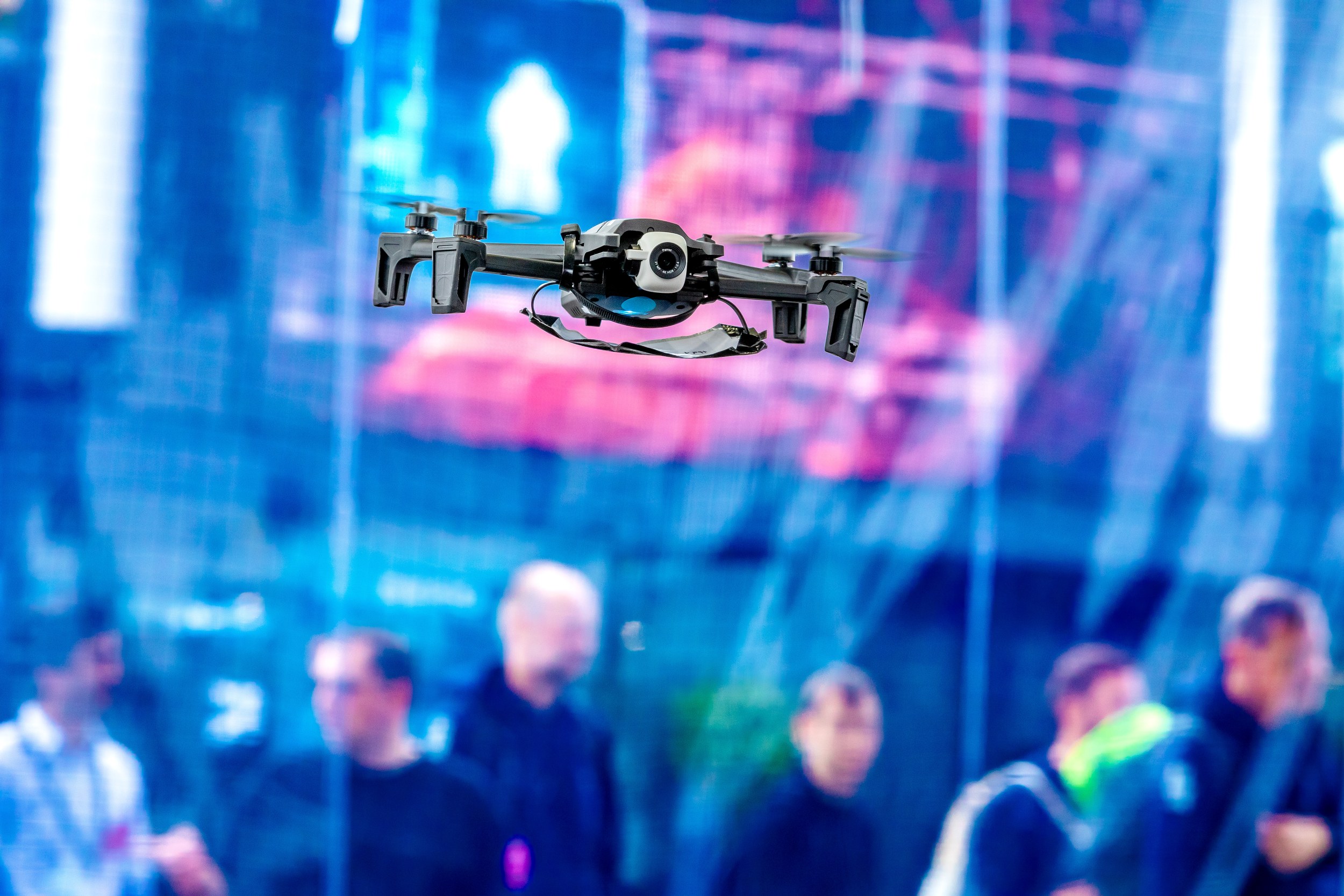 A small black quadcopter hovers head-on against a blue and pink illuminated hall backdrop. Its camera is mounted central