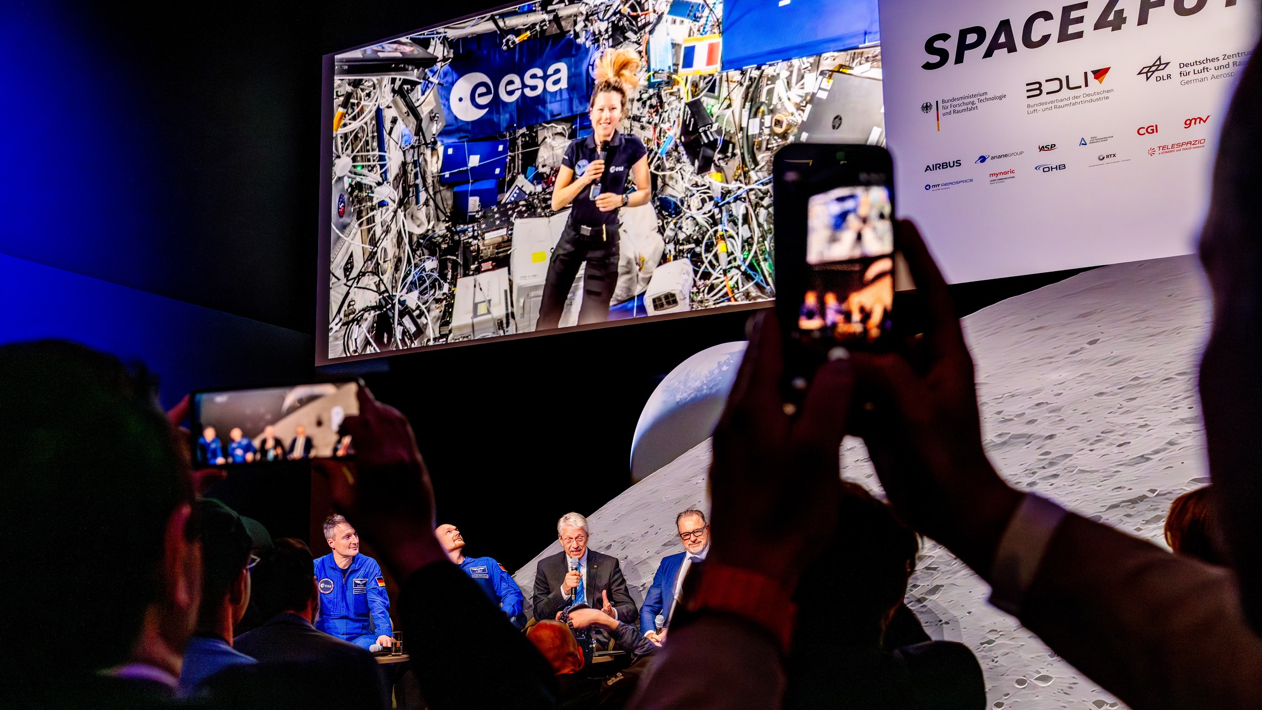 Two raised smartphones frame the view of the ILA STAGE Space as an older panel guest speaks into a handheld microphone. 