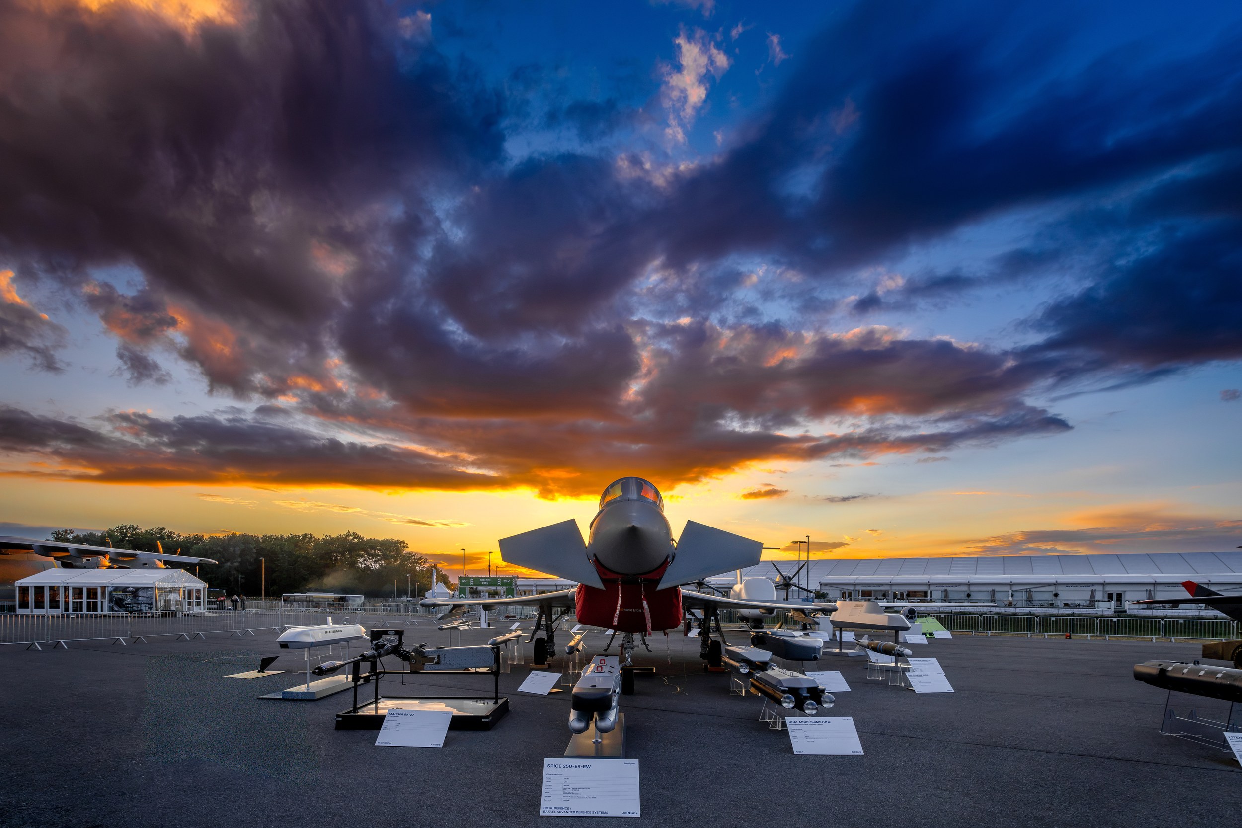 A Eurofighter Typhoon stands head-on in the Defence Park static display beneath a blue and orange evening sky. A long we