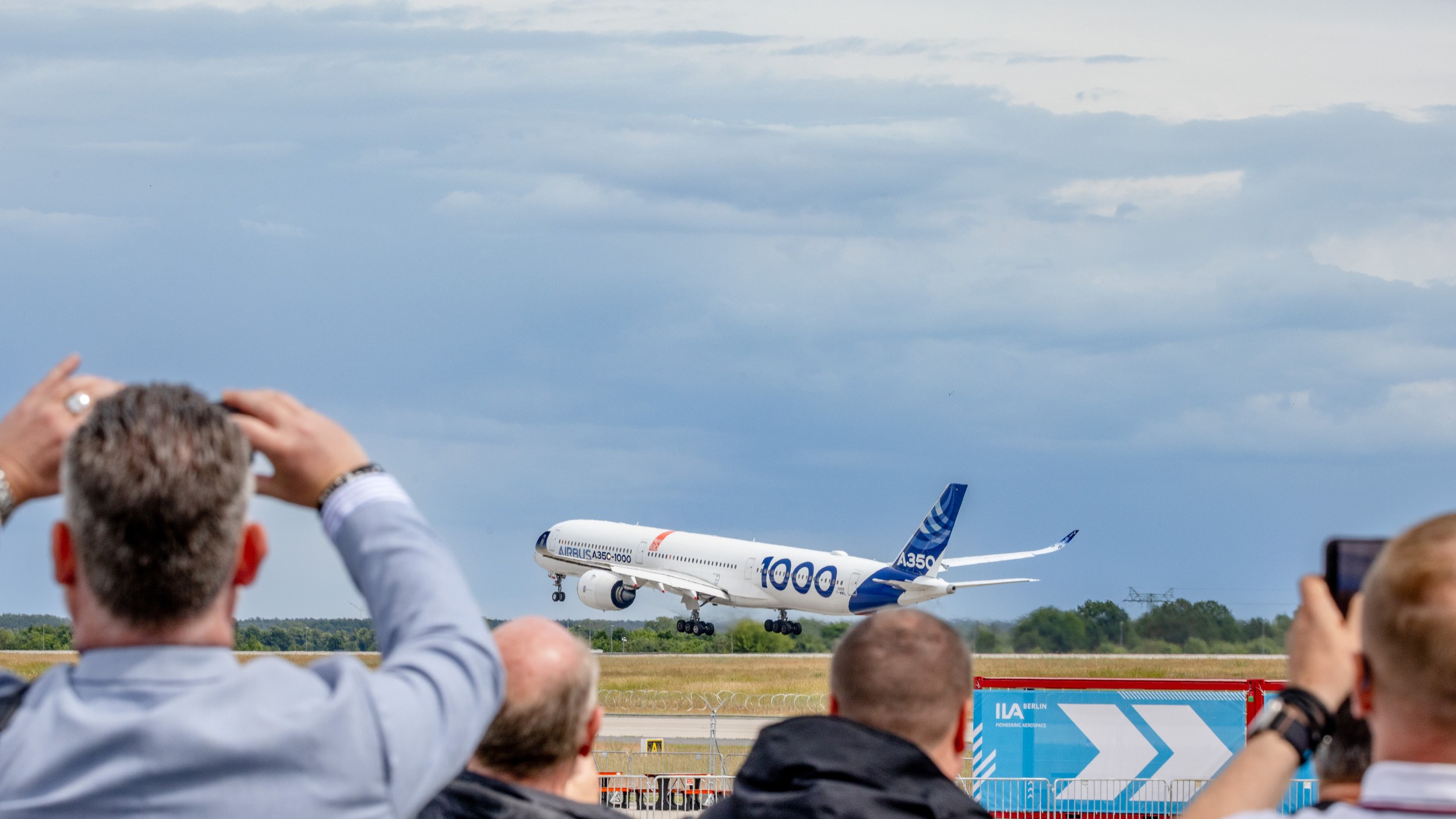 Ein Airbus A350-1000 hebt mit ausgefahrenem Fahrwerk vor einer Reihe fotografierender Zuschauer von der Startbahn ab.