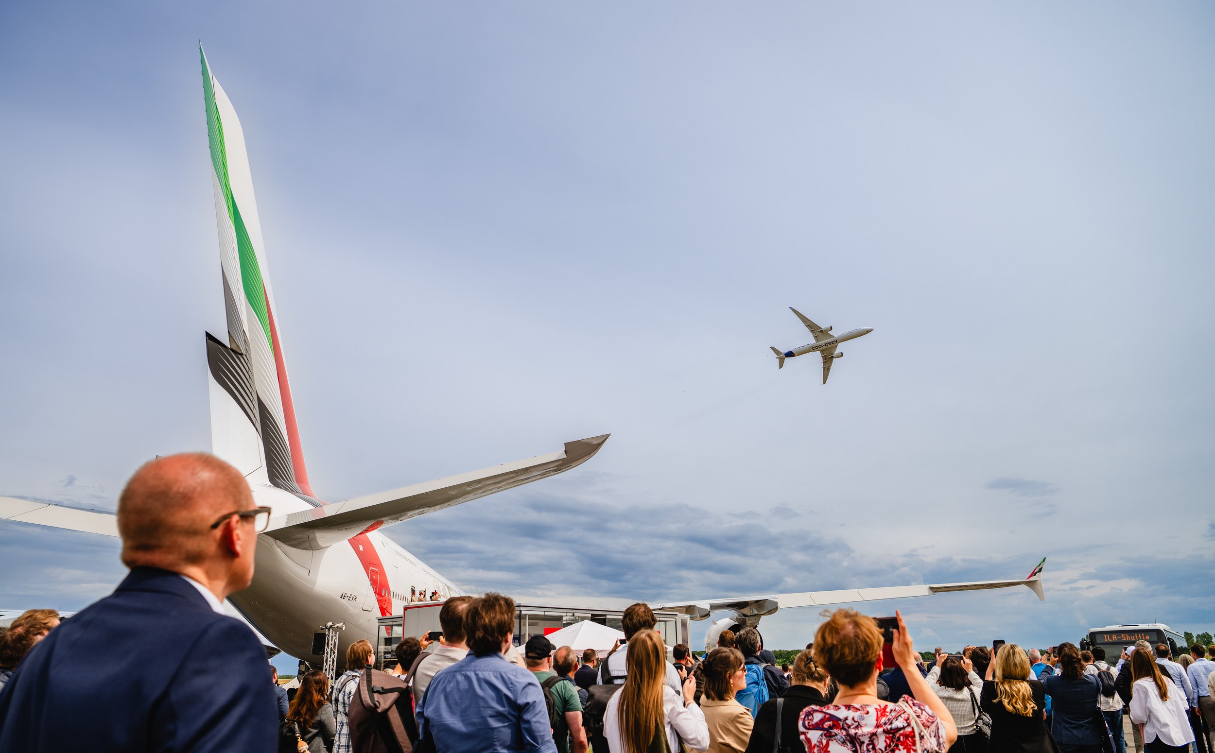 A large visitor crowd watches and photographs an Airbus A350 flying over the outdoor display area.