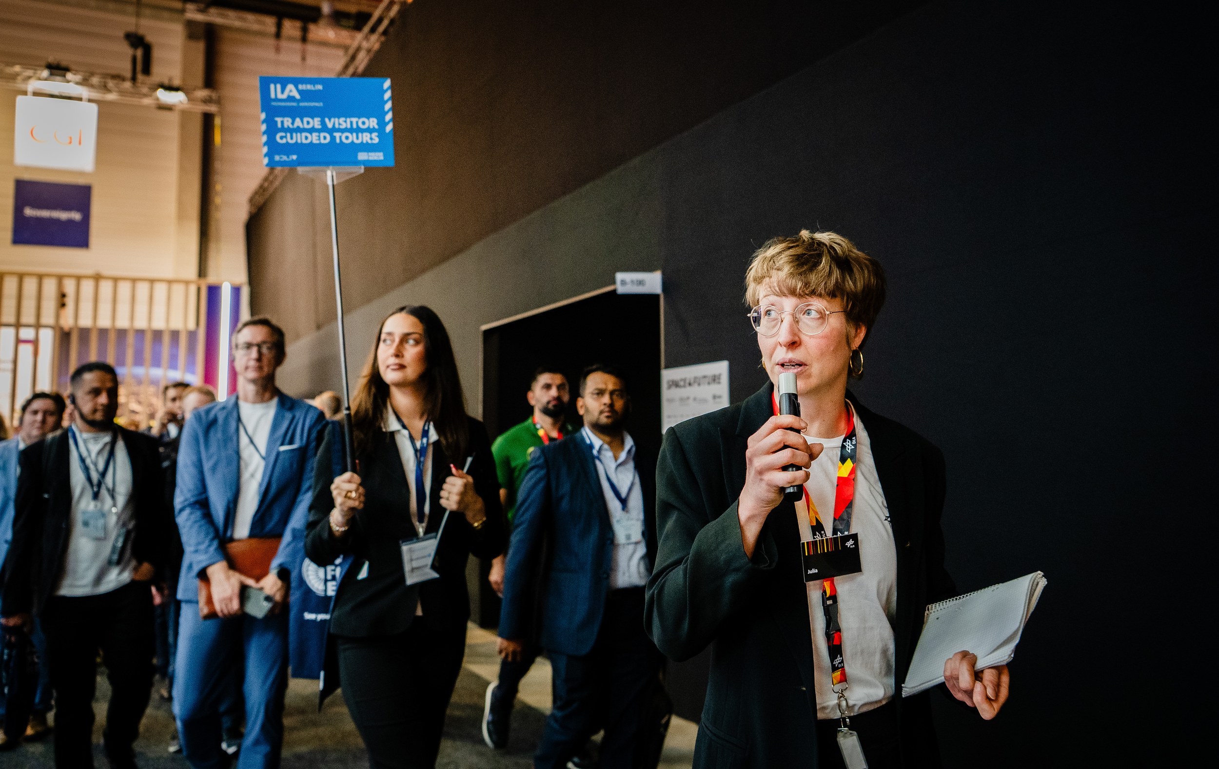 A guide speaks into a microphone and walks ahead of a group of trade visitors through the exhibition hall while carrying a notepad. Another woman holds the blue “TRADE VISITOR GUIDED TOURS” sign as lanyards, badges and the dark aisle frame the tour in the Space Pavilion context.