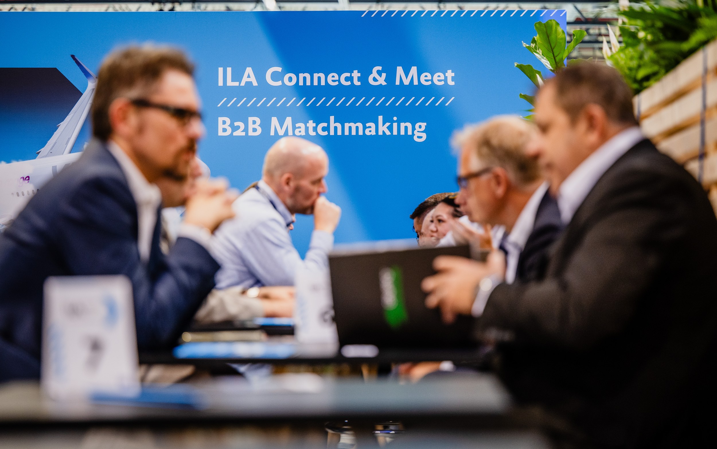 Several trade visitors sit in small discussion groups at tables in the Connect and Meet area, with a laptop and bottles filling the foreground. The people are softened by shallow depth of field, while the blue sign reading ILA Connect & Meet and B2B Matchmaking remains sharp at the centre.