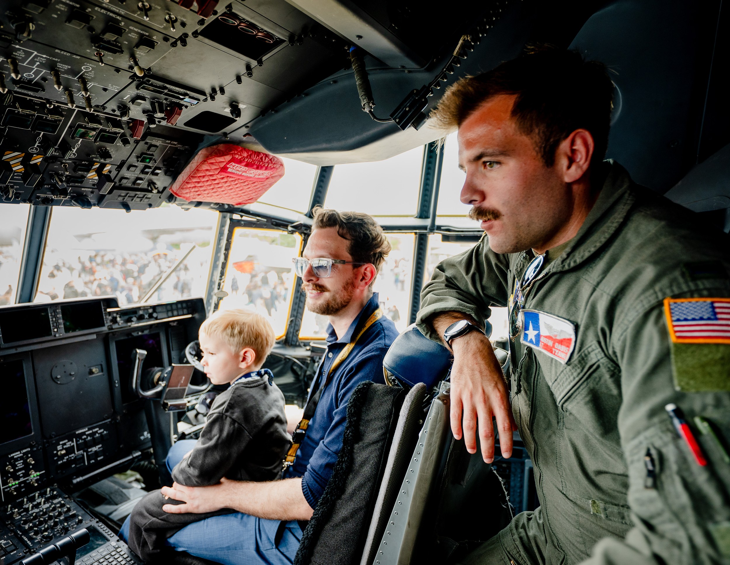 A crew member in a green flight suit leans into the cockpit of a Lockheed Martin C-130 Hercules beside an adult visitor …