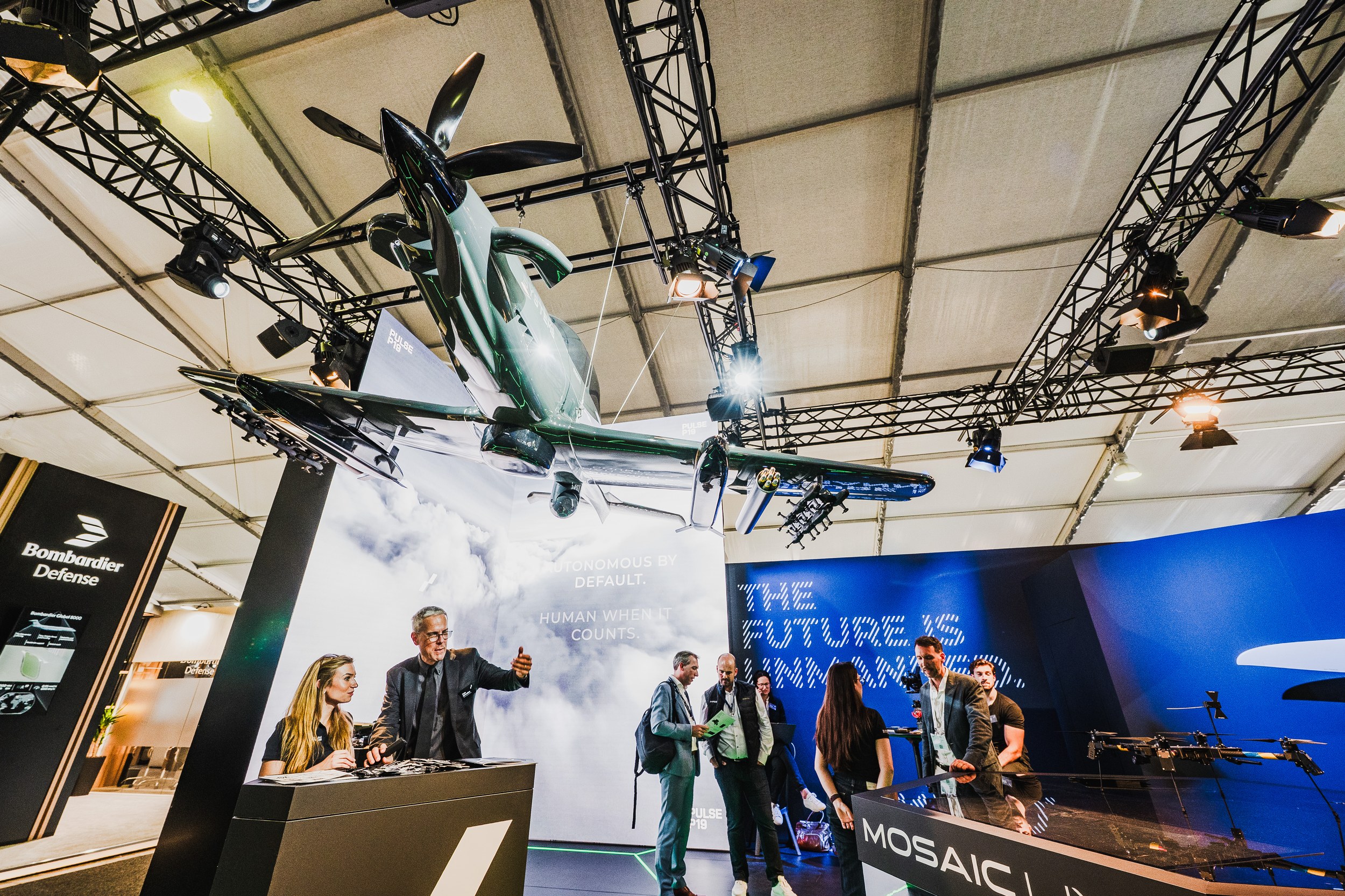A large unmanned aircraft model hangs above the Bombardier Defense stand while trade visitors and booth staff talk below.