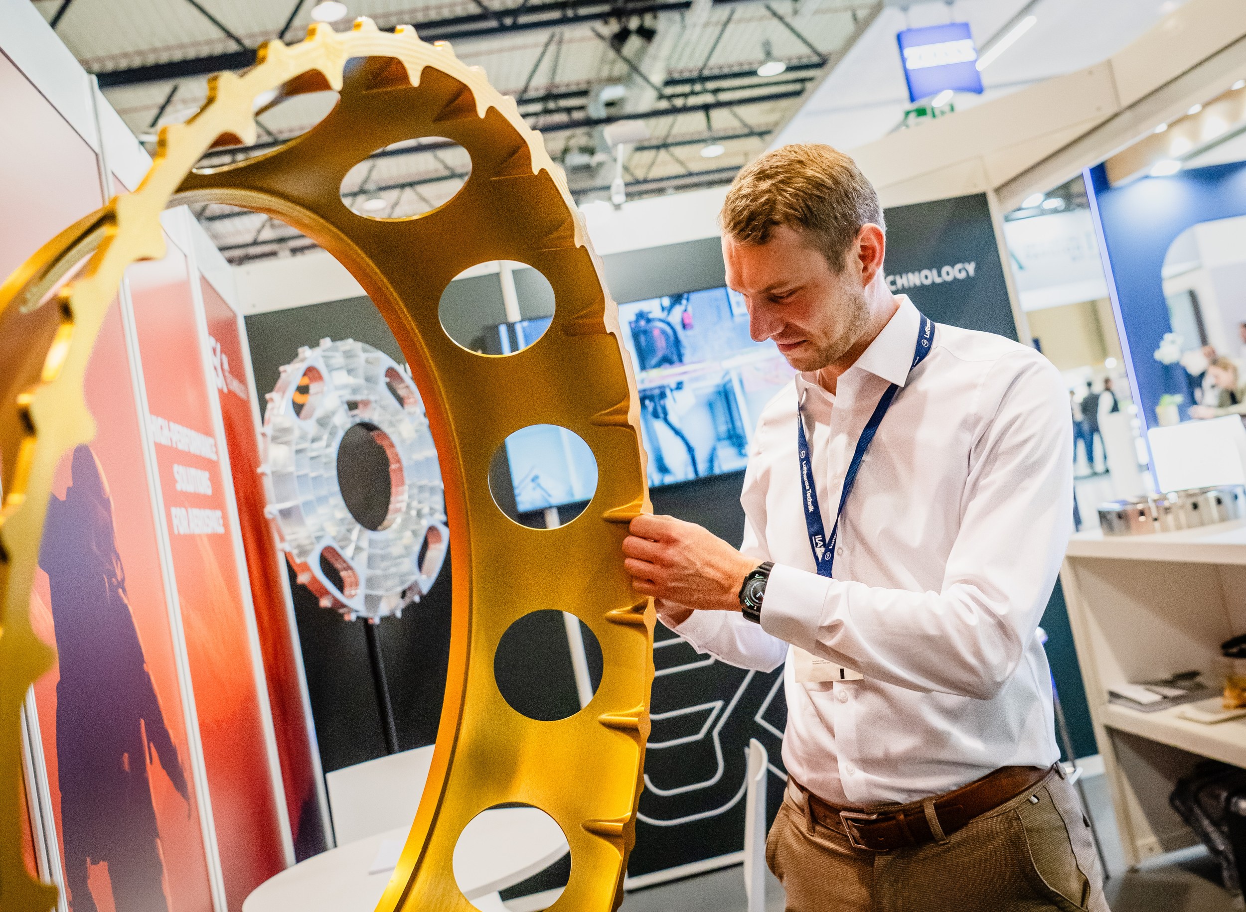 A man in a white shirt uses both hands to inspect a circular opening in the large gold-coloured ring component and studi