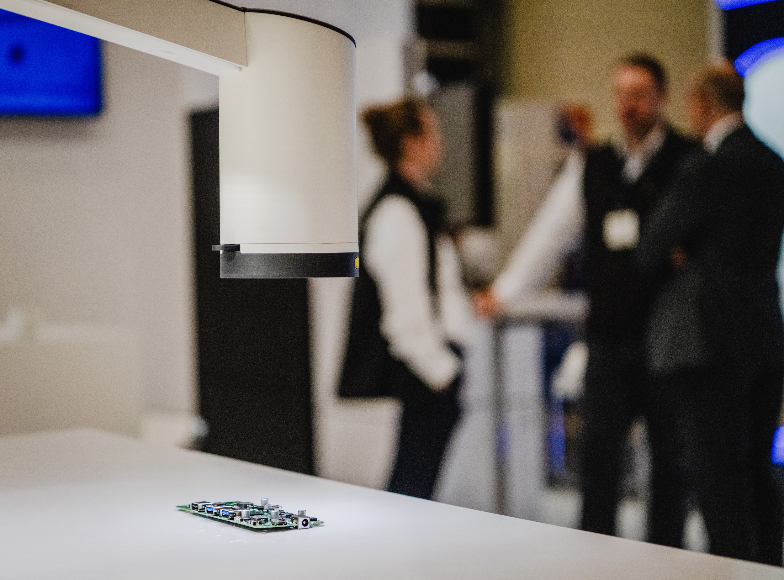 A white technical module with a dark lower edge hangs just above a small populated circuit board on a white tabletop. Several people stand in the heavily blurred exhibition hall background, leaving the possible inspection or scanning device and the board as the visual focus.