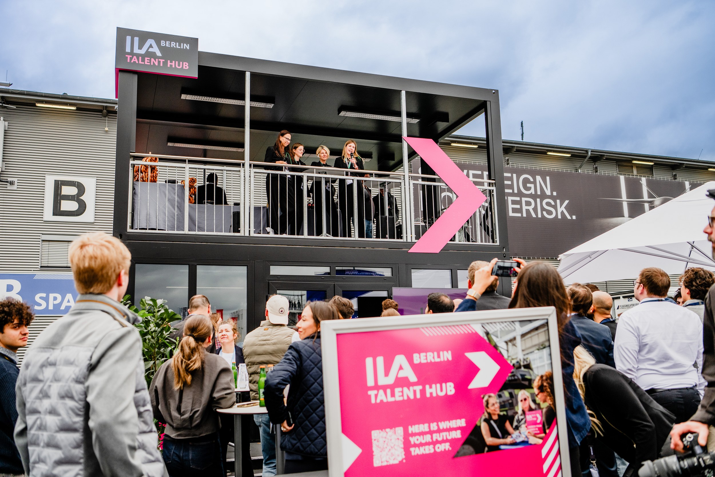 Four speakers stand on the upper platform of the ILA Talent Hub, with one person addressing the crowd below through a microphone. The pink Talent Hub display projects into the foreground of the wide view as a visitor points a camera toward the platform.