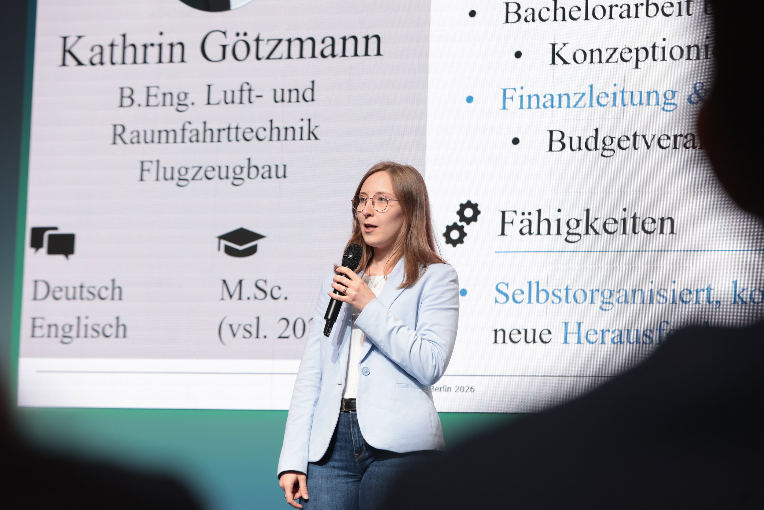 Close stage shot of Kathrin Goetzmann in a light blue blazer speaking into the microphone at the Talent Slam of ILA Berl