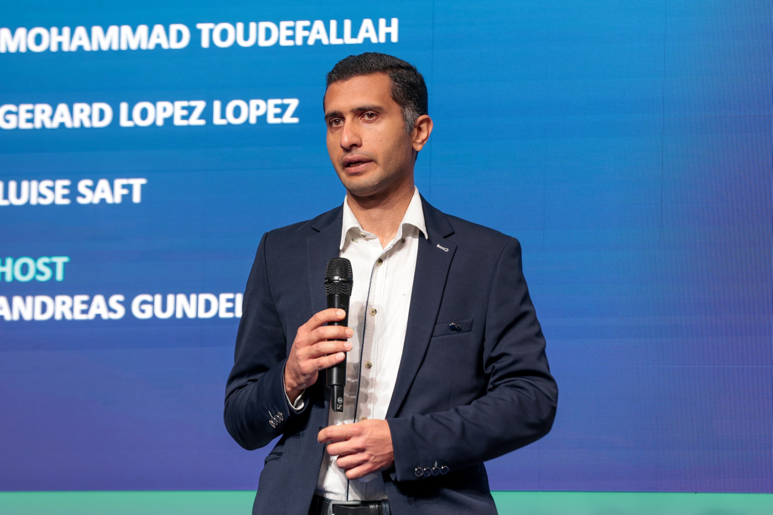 Mohammad Toudefallah stands at the Talent Slam of ILA Berlin 2026 with a microphone in front of a blue screen listing th
